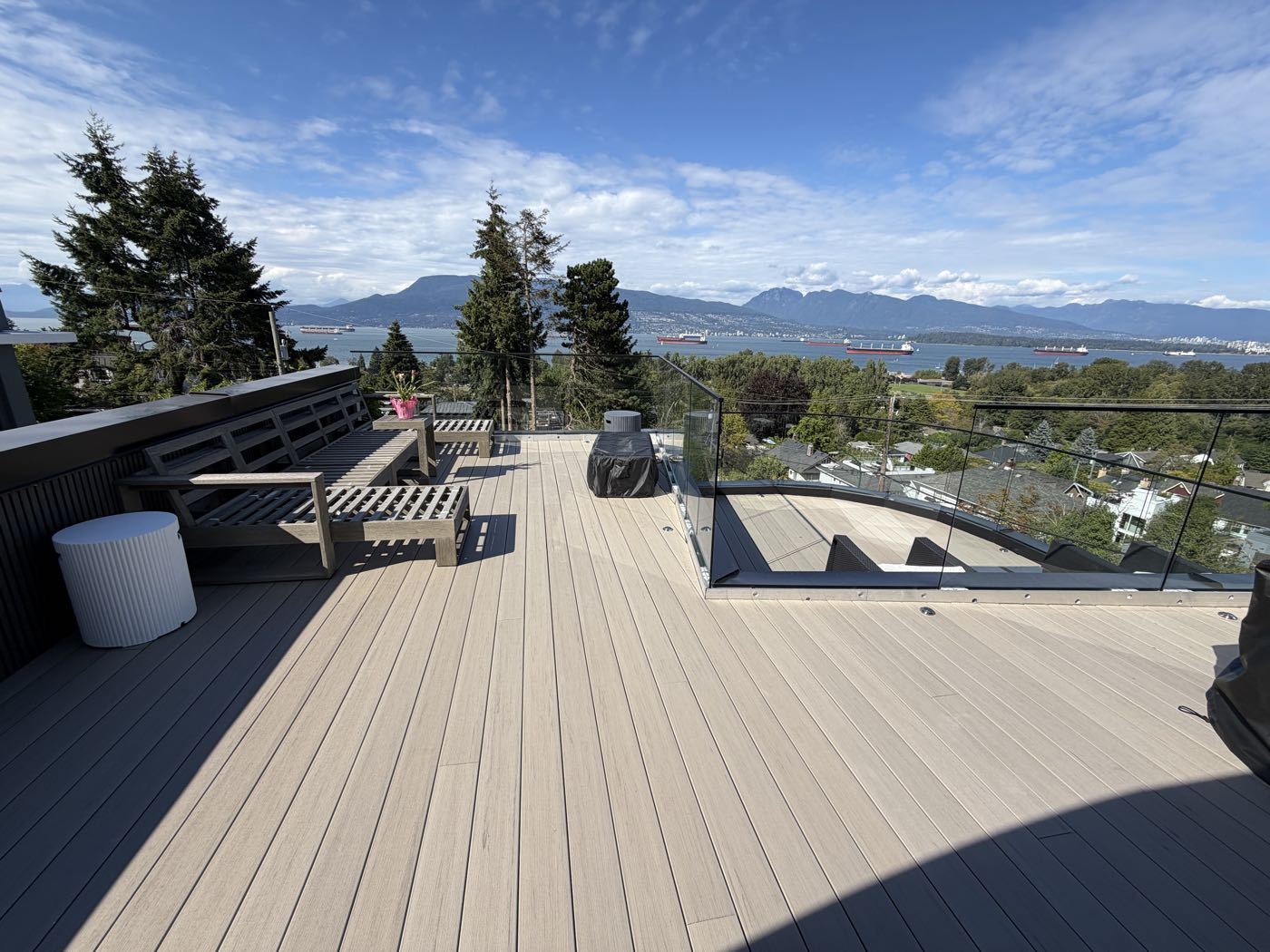 A finished multi-level composite roof deck in Vancouver, looking north over English Bay to the North Shore mountains, with frameless glass guards and a built-in bench along the house wall.